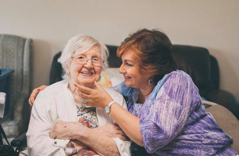 Woman smiling and holding hands with a seated older woman in a living room