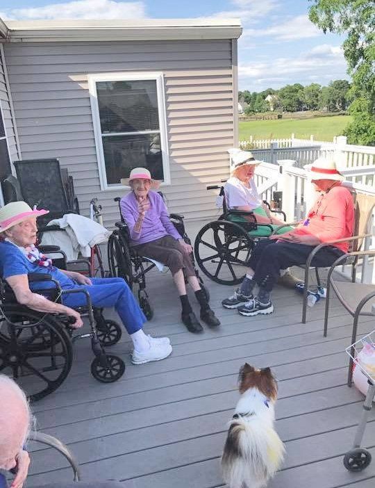 People in wheelchairs and chairs chatting on a sunny patio with a small dog nearby.