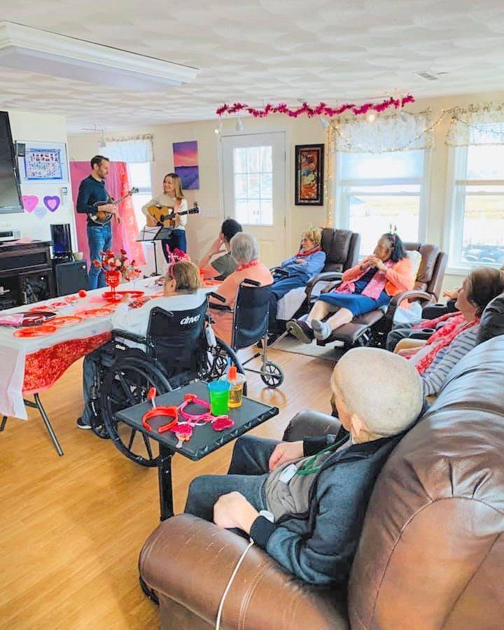 People seated in a bright living room during a holiday gathering, with red streamers and gifts on a table.