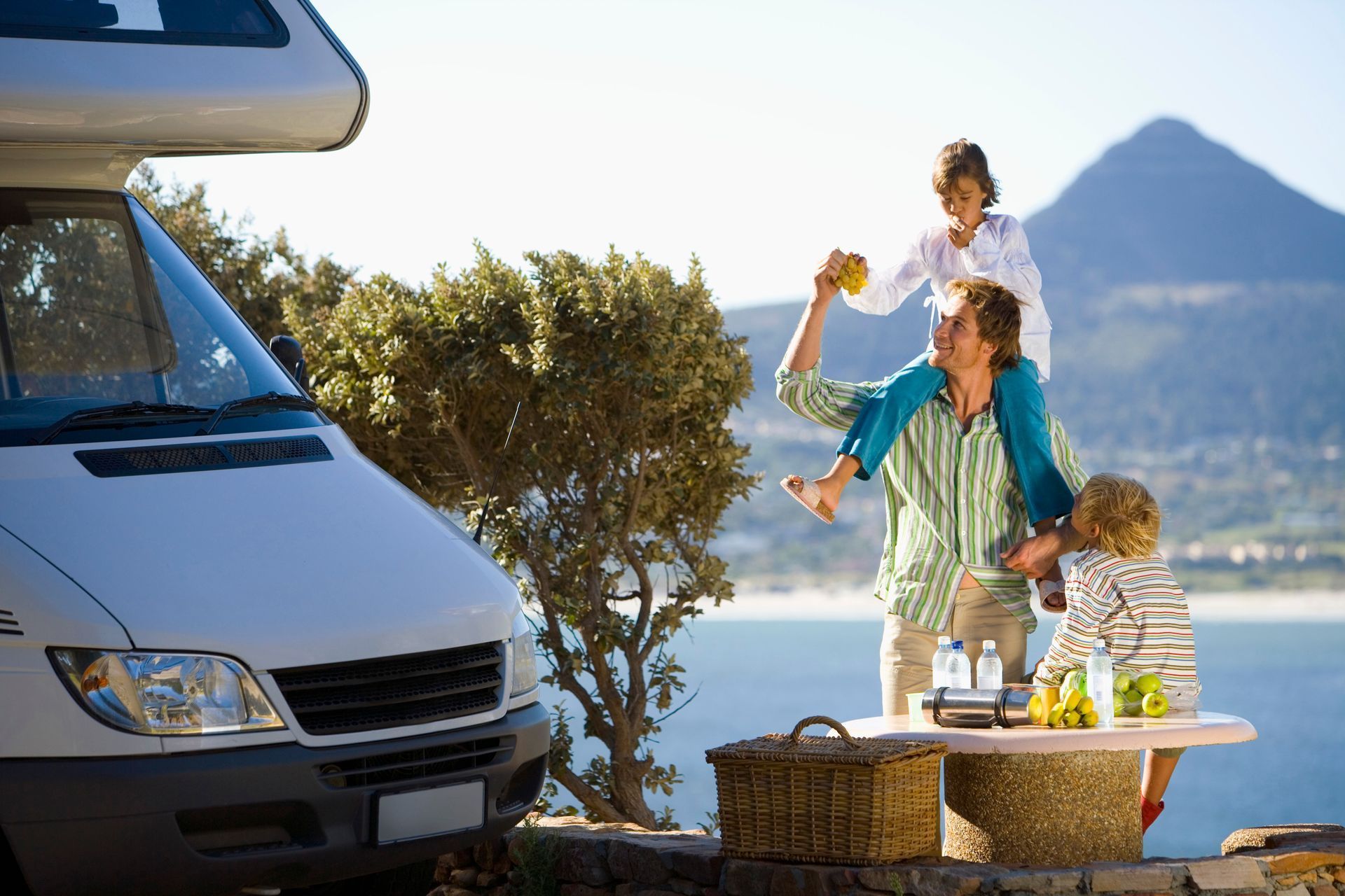 Family beside RV with mountain and water view, picnic table set up.