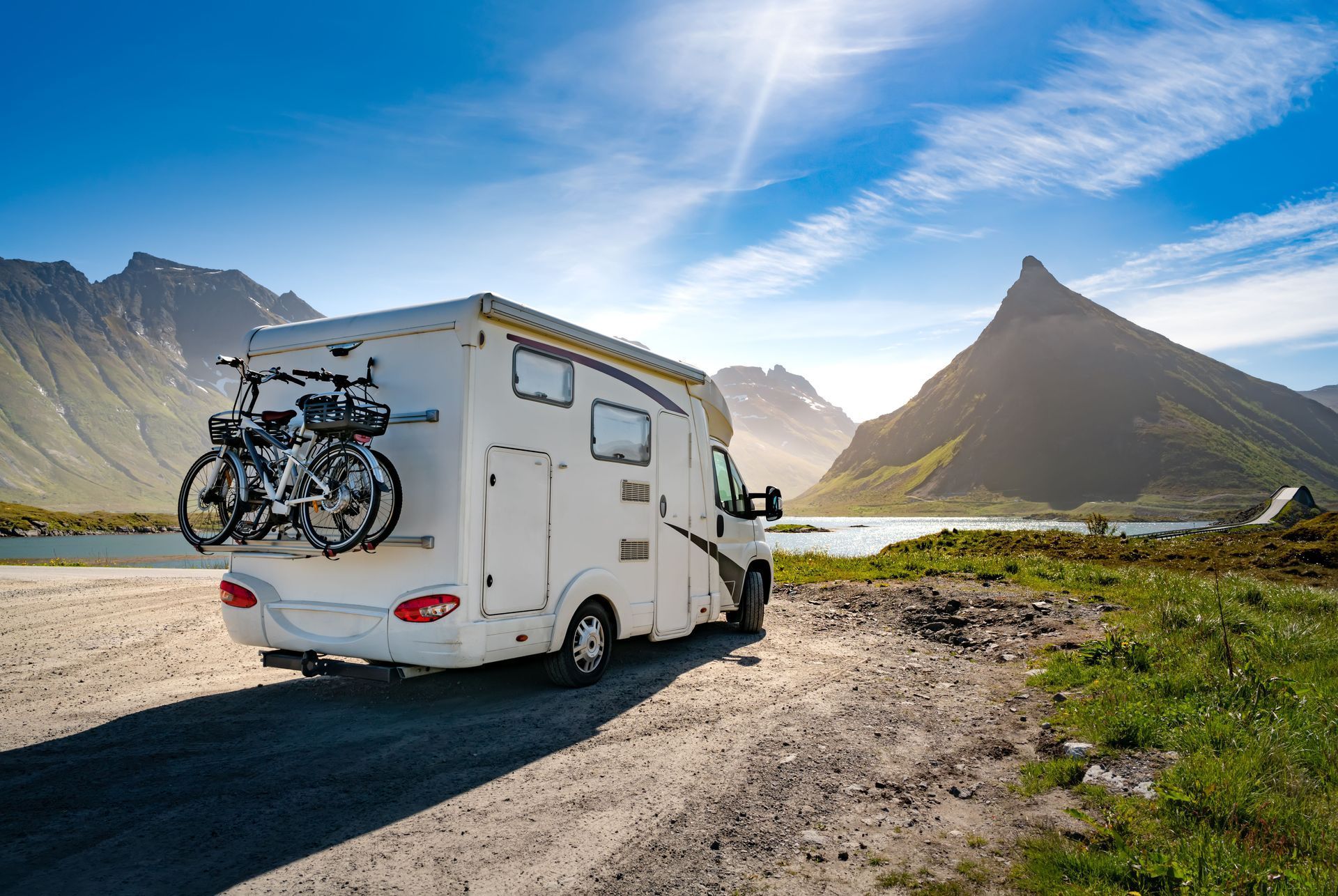 White RV parked on a dirt road by a lake, mountains in background, sunny day.