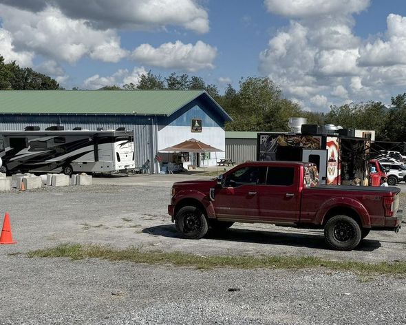 Red pickup truck parked in front of a blue and white building with an RV and gravel lot.