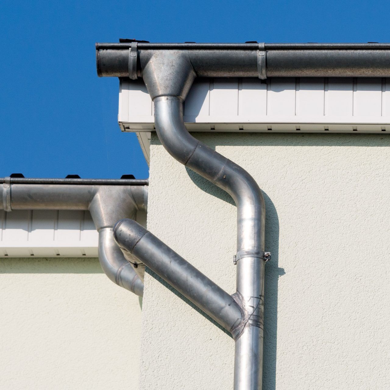 Silver rain gutters and downspouts on a light-colored house against a blue sky.