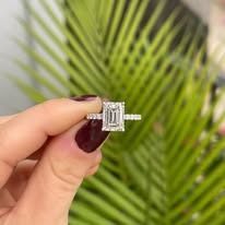 Hand holding a diamond emerald-cut ring in front of green leaves.