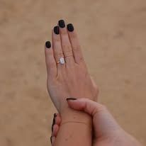 Hand with black nail polish, wearing an emerald-cut diamond engagement ring, resting on another hand.
