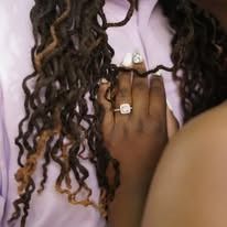Person's hand wearing diamond ring, resting on a chest. Brown dreadlocks frame the person, wearing a pink shirt.