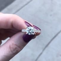 A hand holding a diamond engagement ring with a large central stone and smaller band stones.