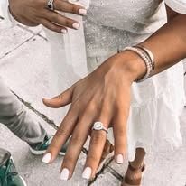 Woman's hands with engagement ring and bracelets; white dress, outdoor setting.