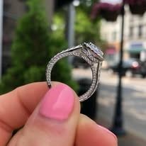 Woman's hand holding a diamond engagement ring, pink nail polish, outdoor setting.