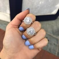 Hand wearing two silver rings with round, diamond-studded halos; light blue nail polish.