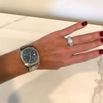 Person's hand with watch and ring. Hand is on a white surface. Red nail polish.