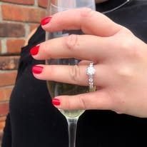 Woman with red nails holding champagne, showcasing a diamond engagement ring.