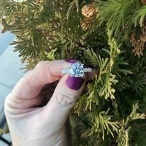 A hand with a purple nail holds a large, sparkling diamond ring near green foliage.