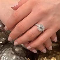 Person's hand wearing a diamond ring with a halo setting, resting on a reflective surface.