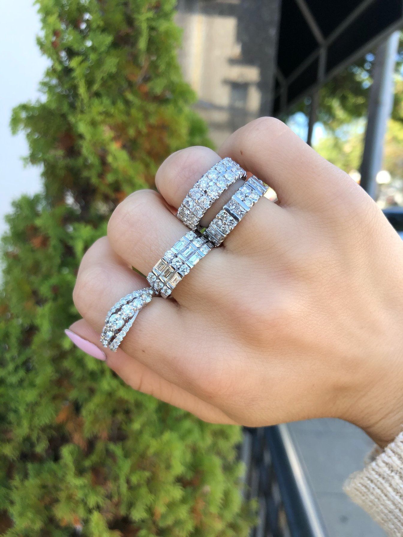 Hand wearing multiple diamond-encrusted rings against a green bush background.