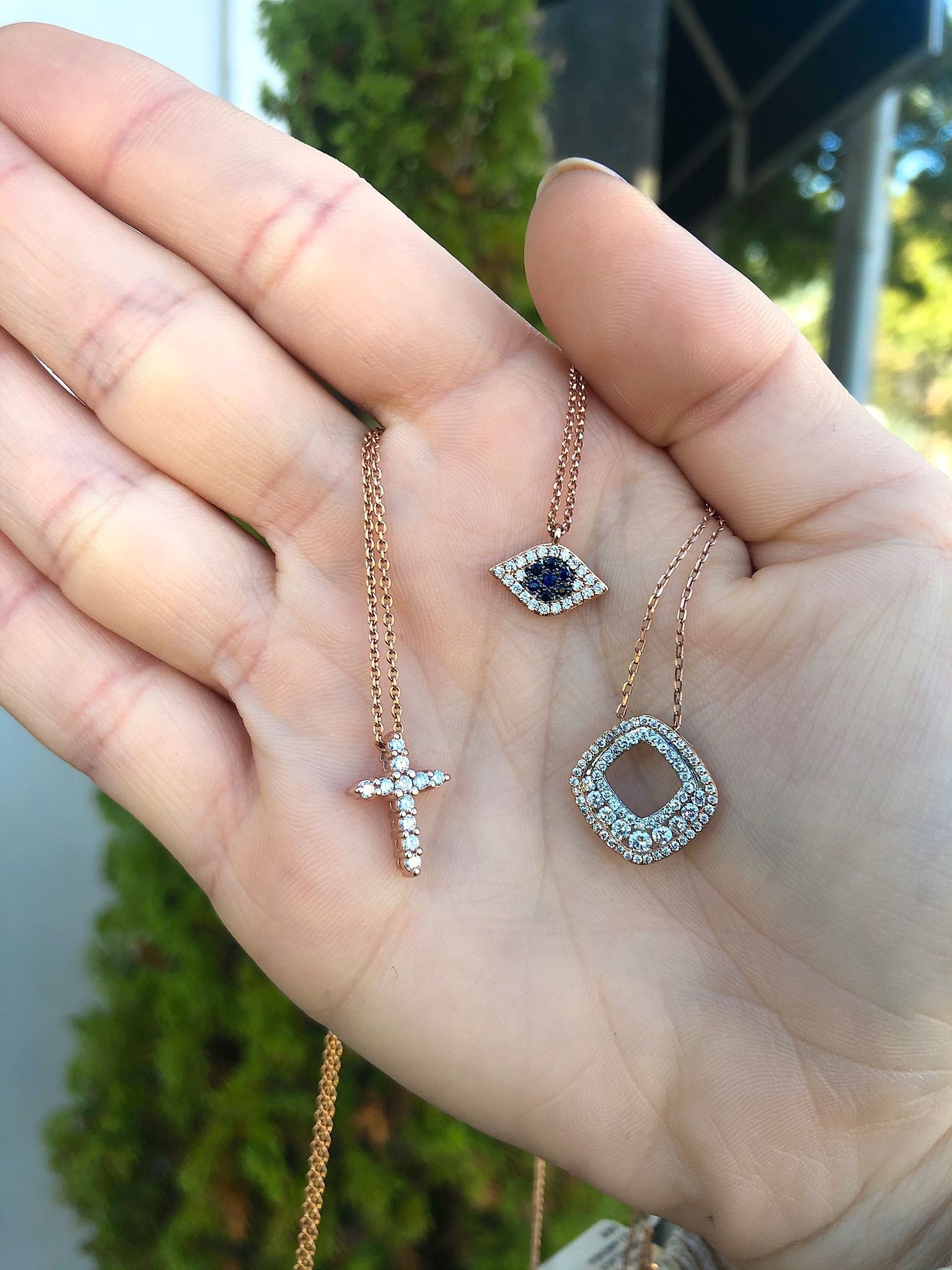 Hand holding three rose gold necklaces: cross, eye, and flower pendant.