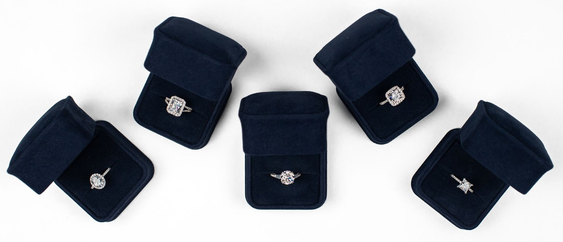 Six dark blue velvet ring boxes, each containing a diamond engagement ring, sit on a white surface.
