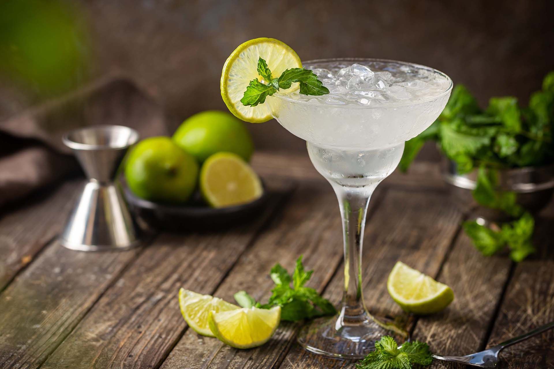 Martini glass with a lime garnish on a rustic table, beside limes, mint, and a cocktail shaker