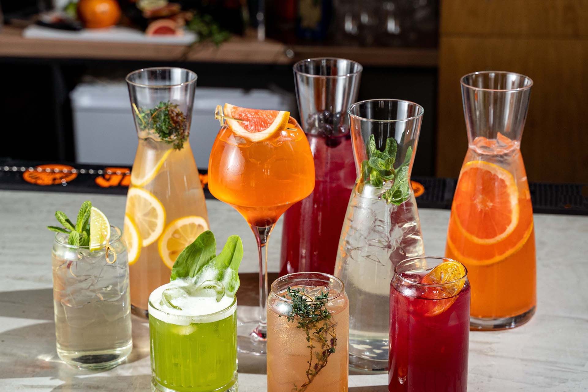 Assorted colorful cocktails and mocktails in tall glasses with garnishes on a bar counter.