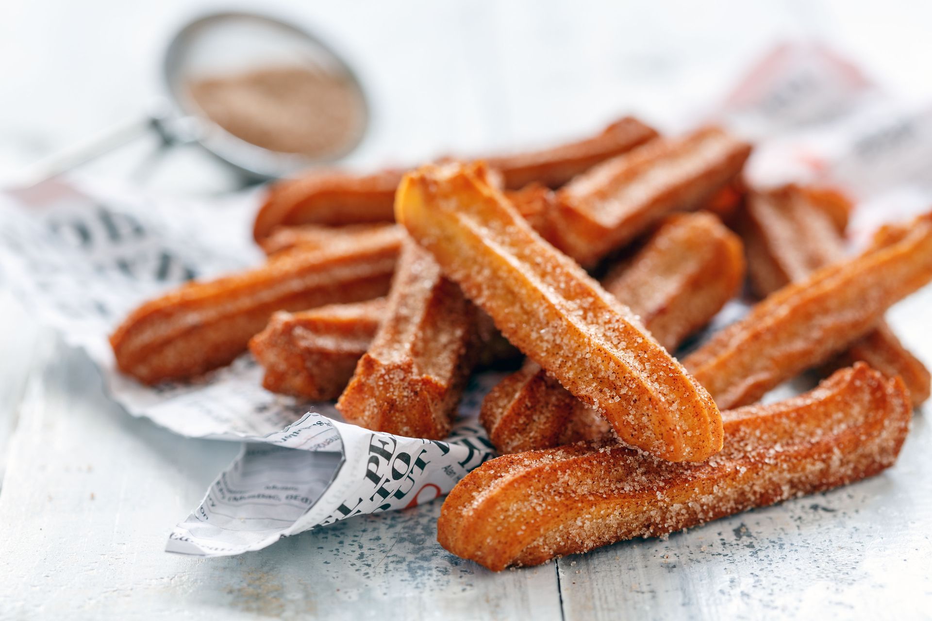 Golden churros dusted with sugar on white paper beside a small cup of dipping sauce