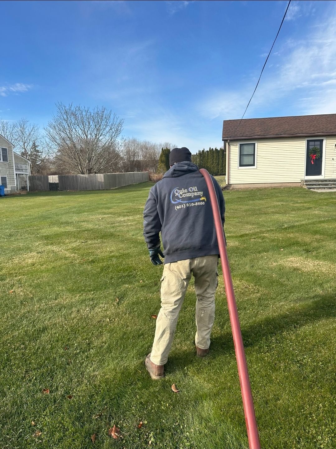 A person in a gray hooded sweatshirt and tan pants walks across a grassy yard while holding a long, red service hose.