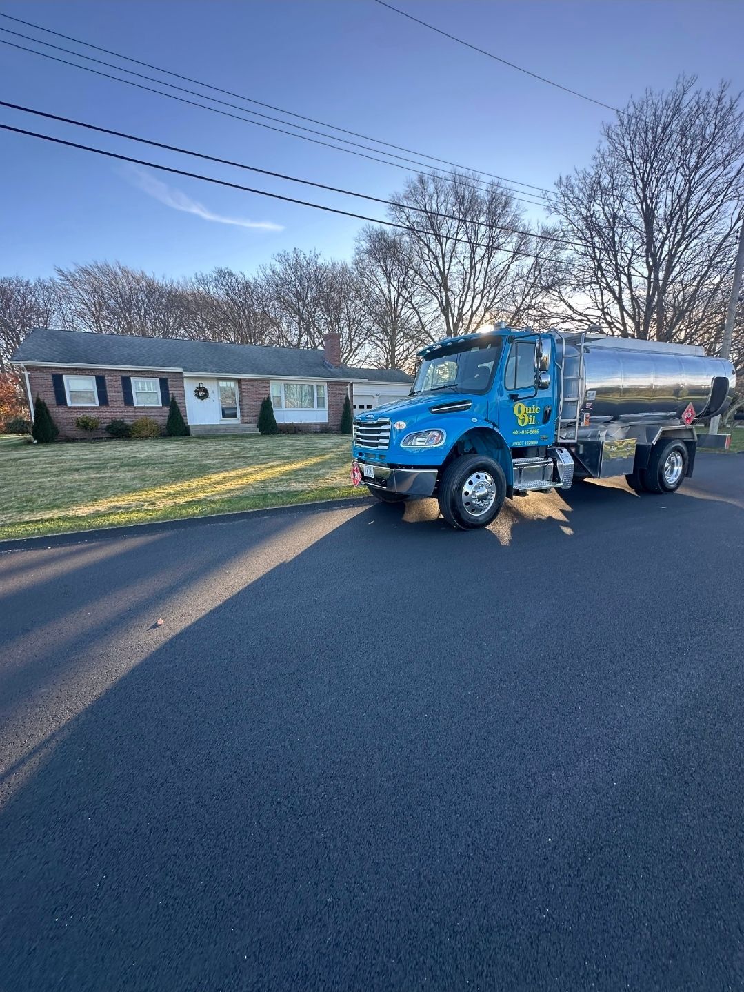 A blue oil delivery truck parked on a residential street in front of a brick house on a sunny day.