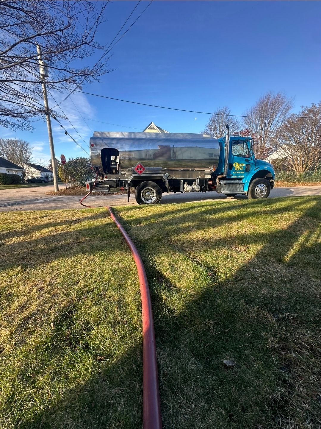 A teal fuel delivery truck with a chrome tank parked on the side of a road, with a long red hose extending onto the grass.