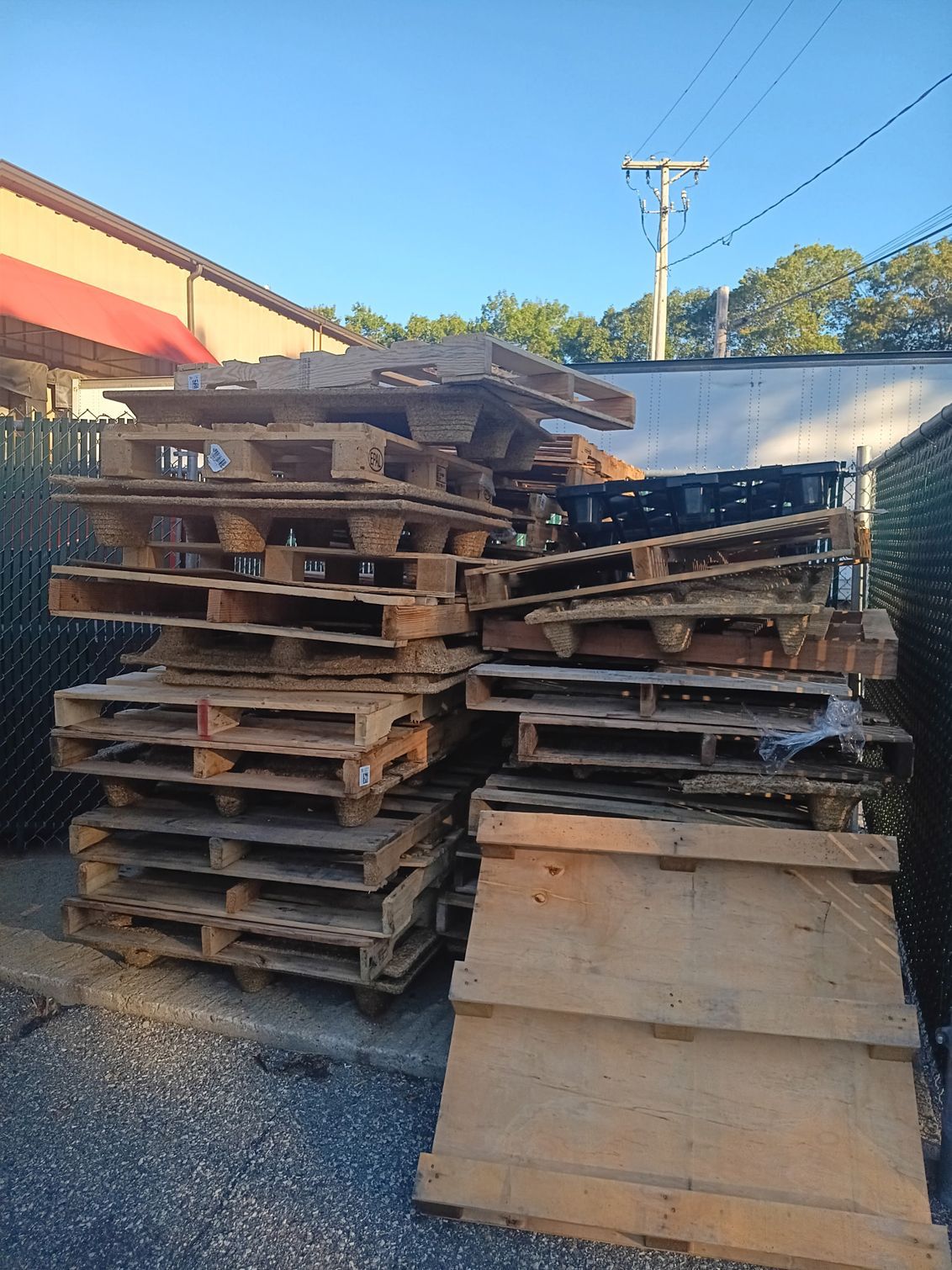 Pile of wooden pallets stacked outdoors, against a fence and building, under a blue sky.