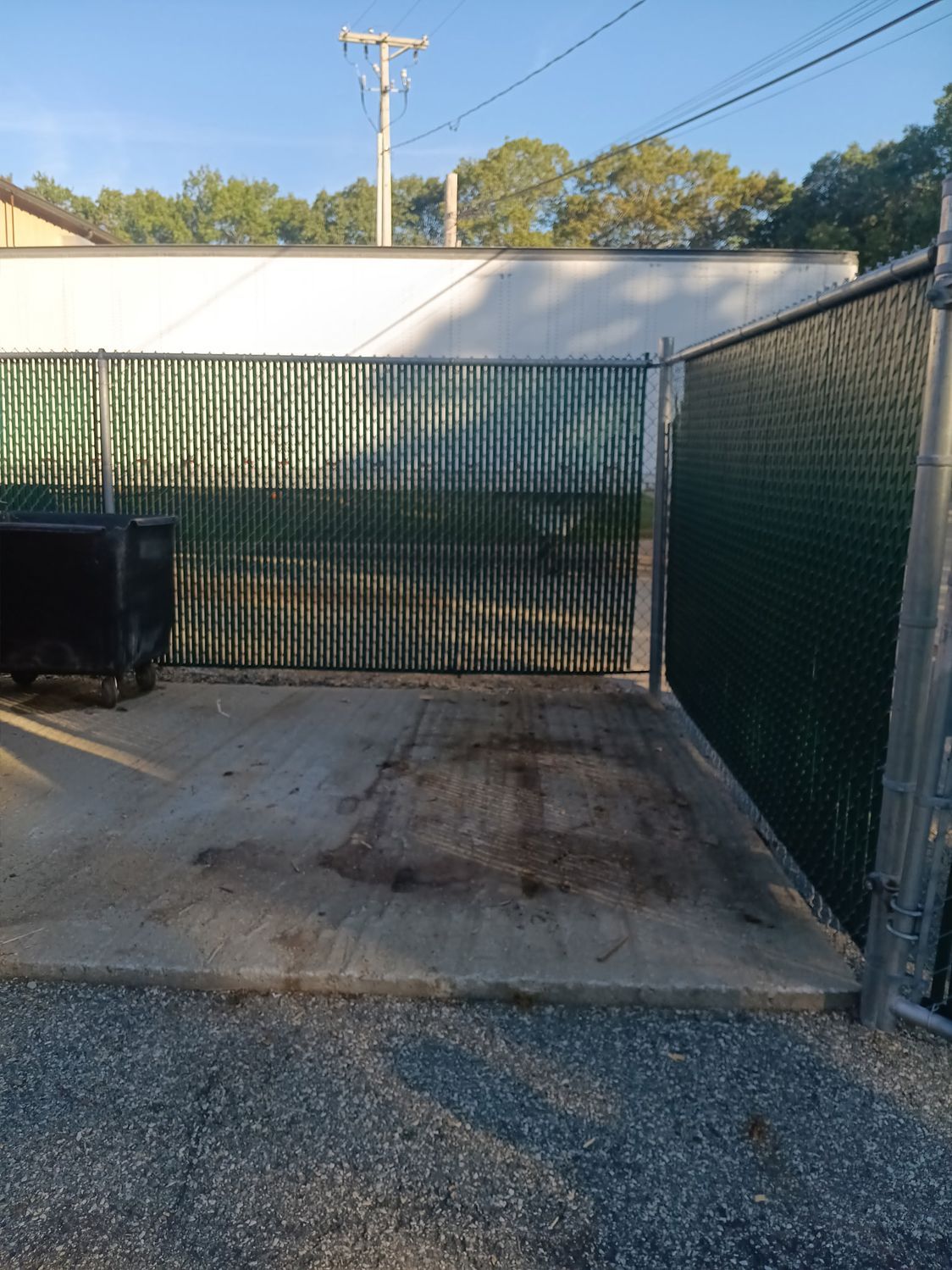Concrete pad with a chain-link fence, a dumpster, and a white building in the background.