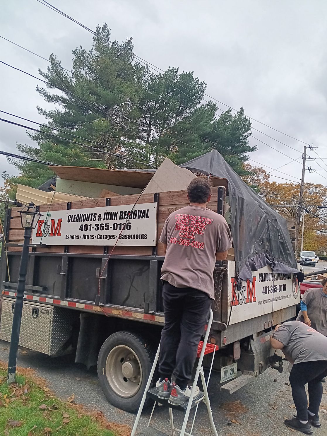 People loading a truck with construction debris; one on a ladder. Overcast day.