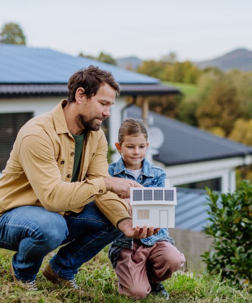 A man and a child are kneeling in the grass holding a model house.