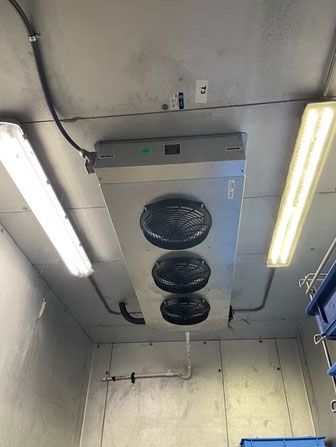 Ceiling-mounted industrial ventilation unit with three fans in a utility room, lit by fluorescent lights