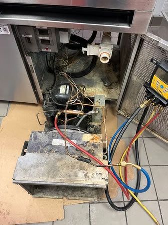 Open refrigerator base with exposed wiring, compressor, and cleaning tools on the floor