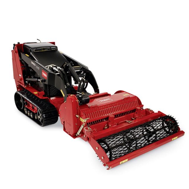 Red Toro Dingo compact utility loader with a soil cultivator attachment. Tracks and controls visible.