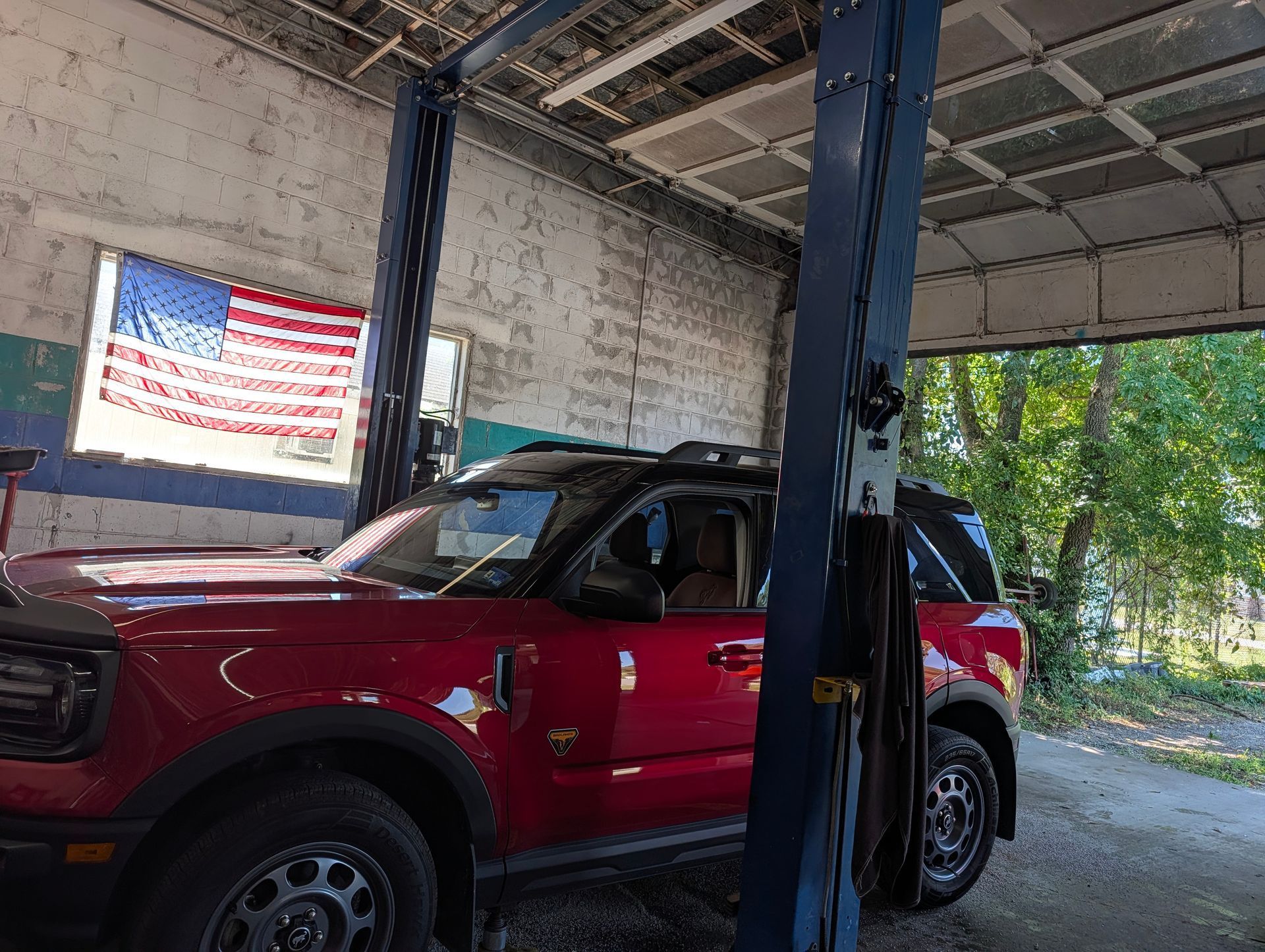 Red SUV parked inside an auto repair garage under a lift, with an American flag banner on the wall.