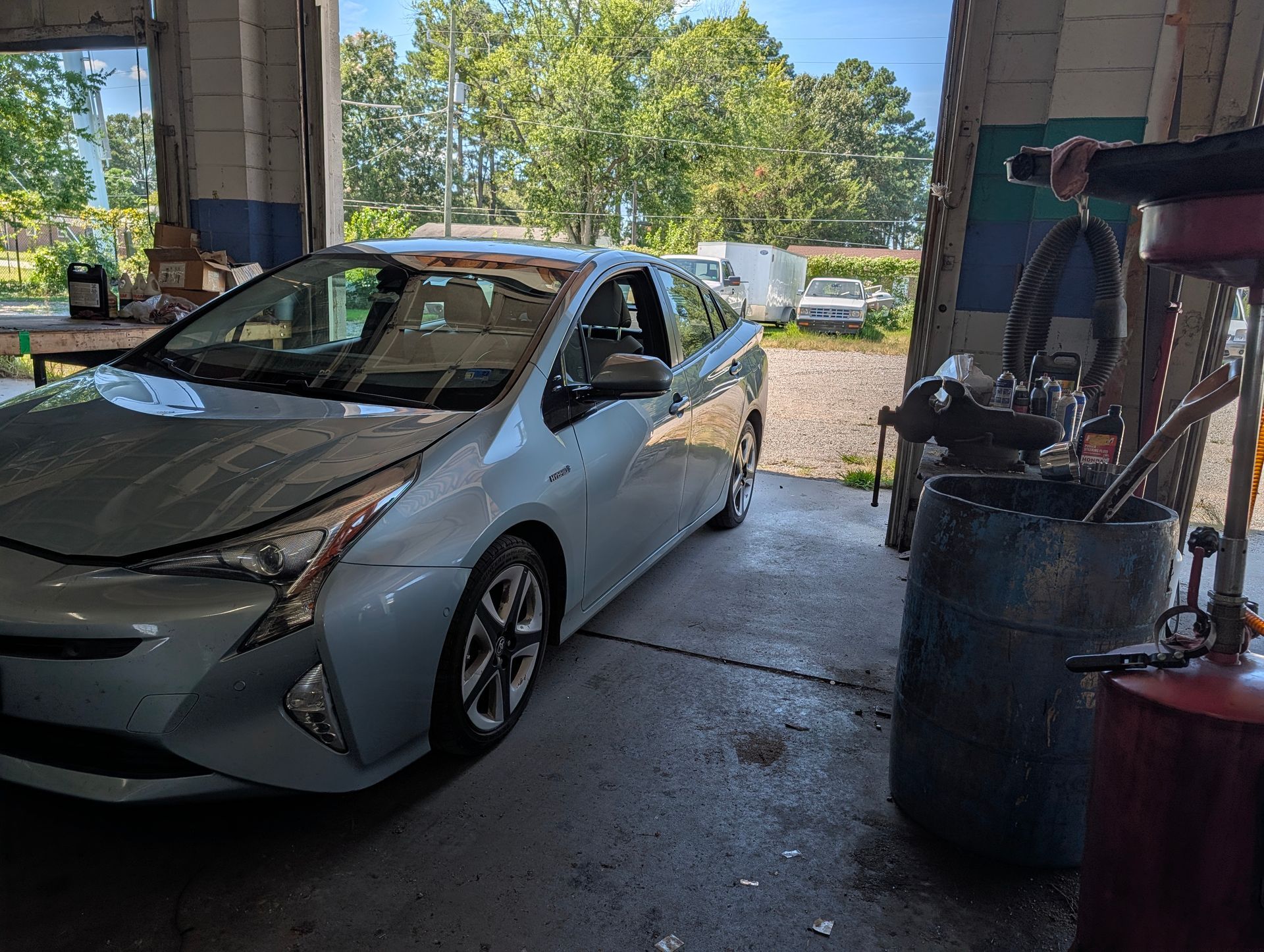 Silver sedan parked in a garage beside welding equipment and barrels, with a sunny outdoor view beyond.