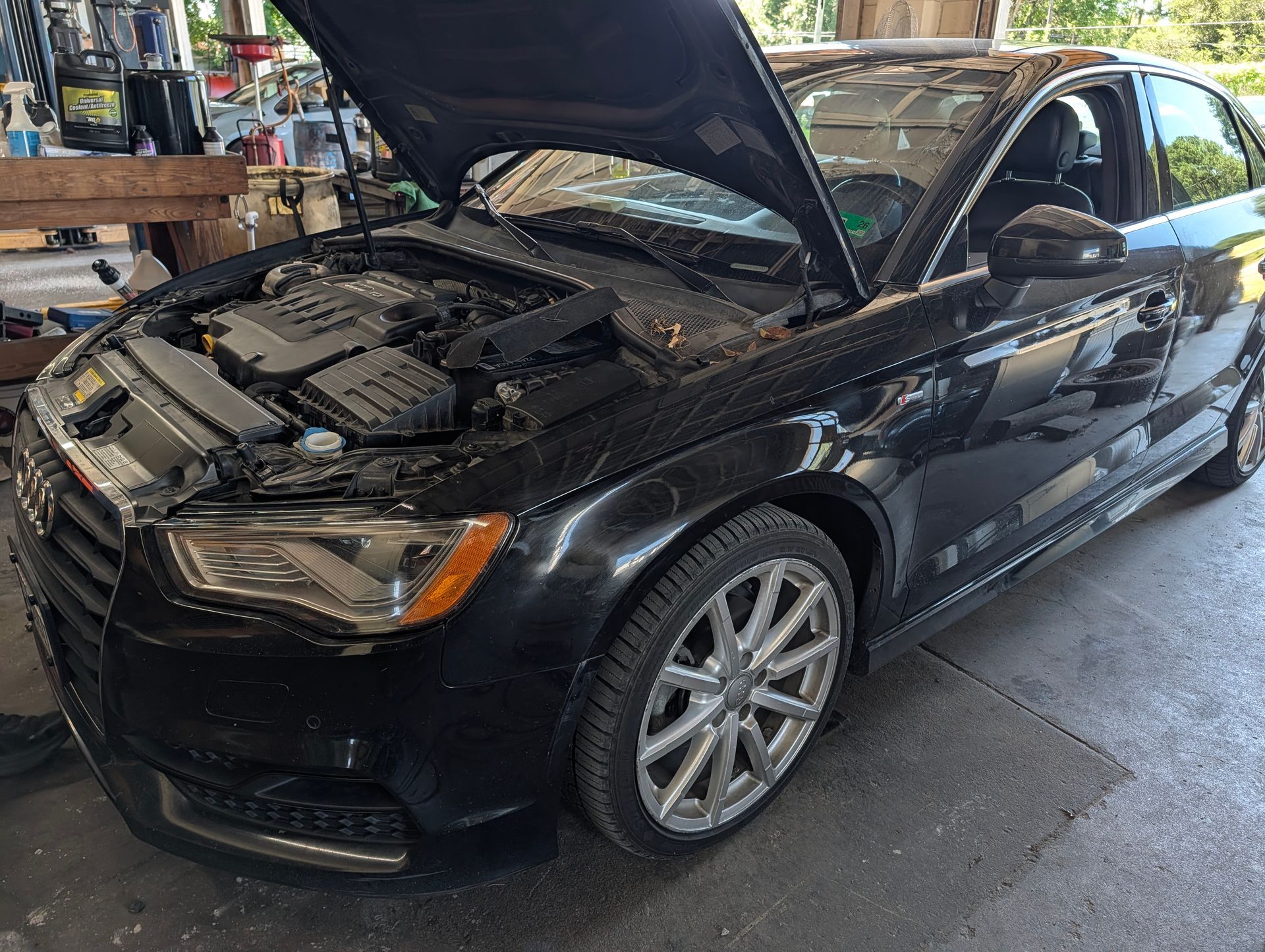 Black sedan with hood open in a garage, showing engine bay and front wheel.