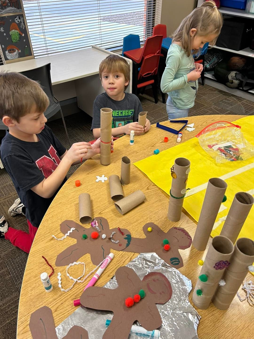 A group of children are sitting at a table making gingerbread men out of toilet paper rolls.