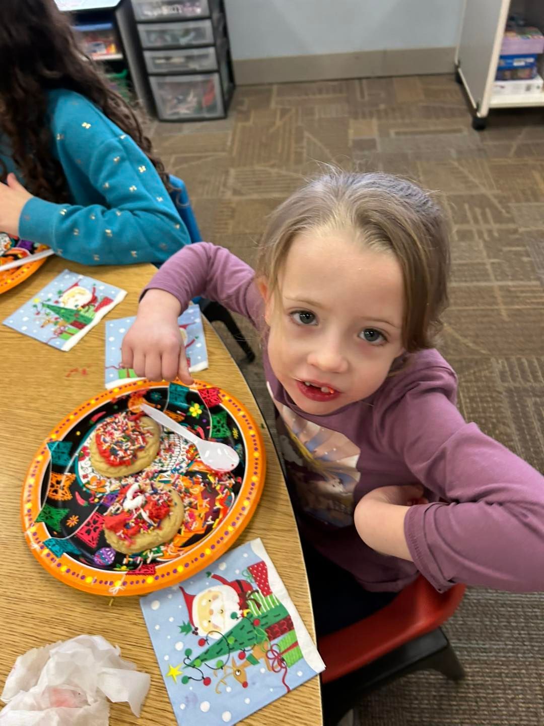 A little girl is sitting at a table with a plate of cookies on it.