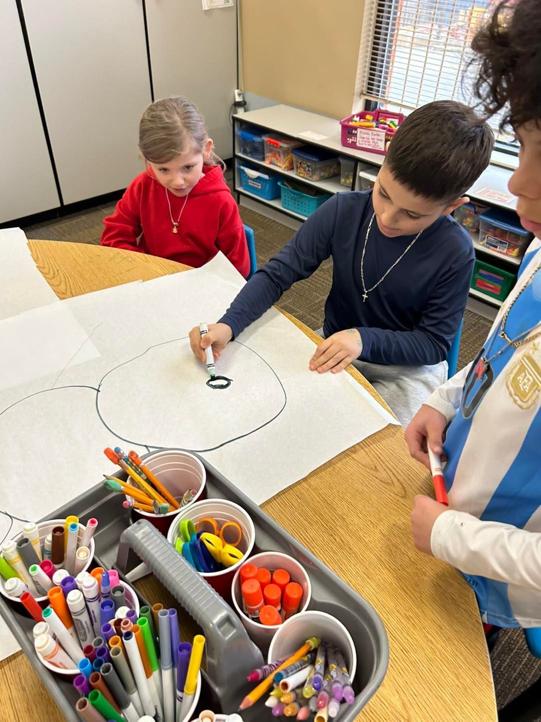 A group of children are sitting at a table drawing on a piece of paper.