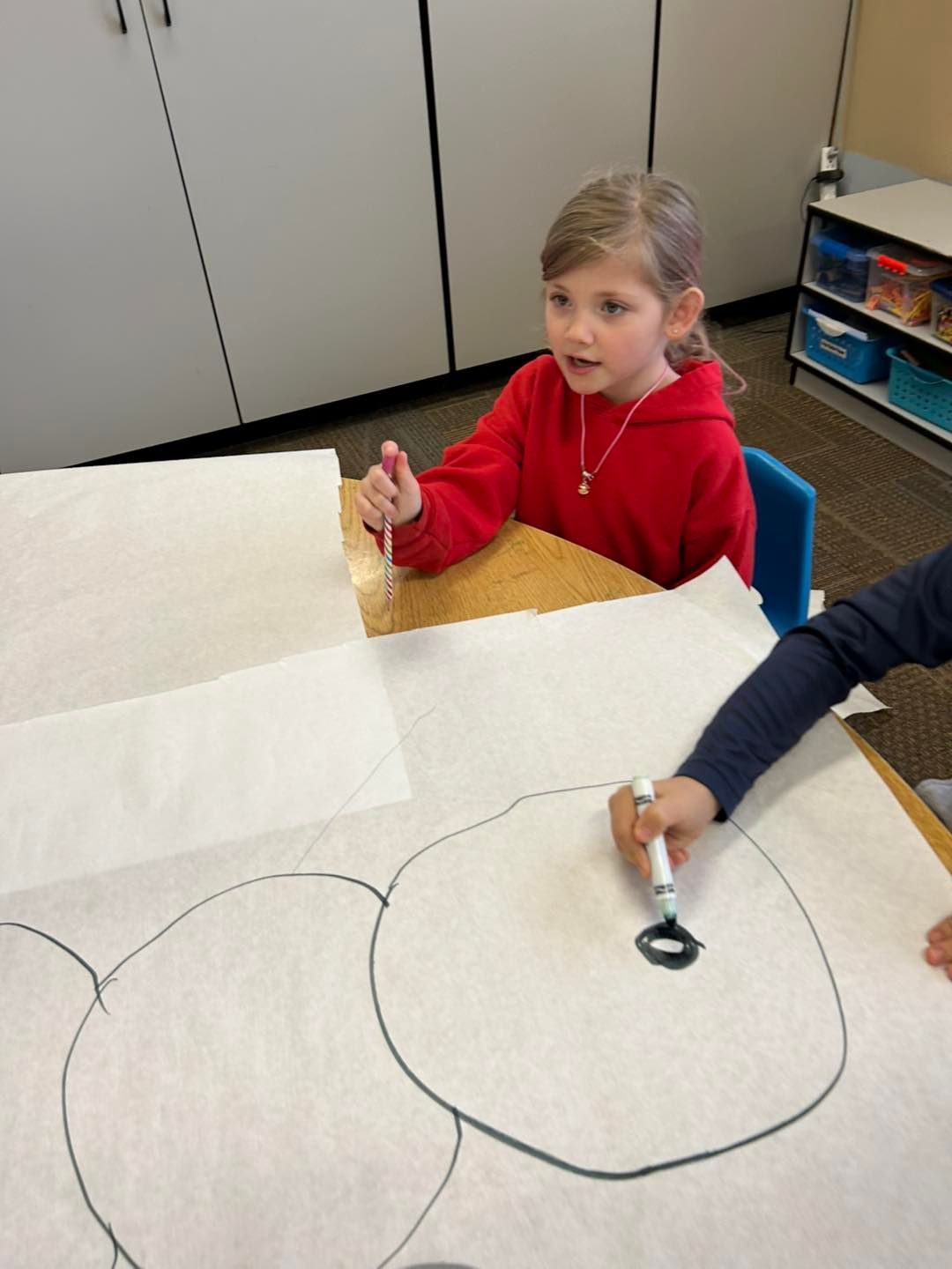 A little girl is sitting at a table drawing a bear with a marker