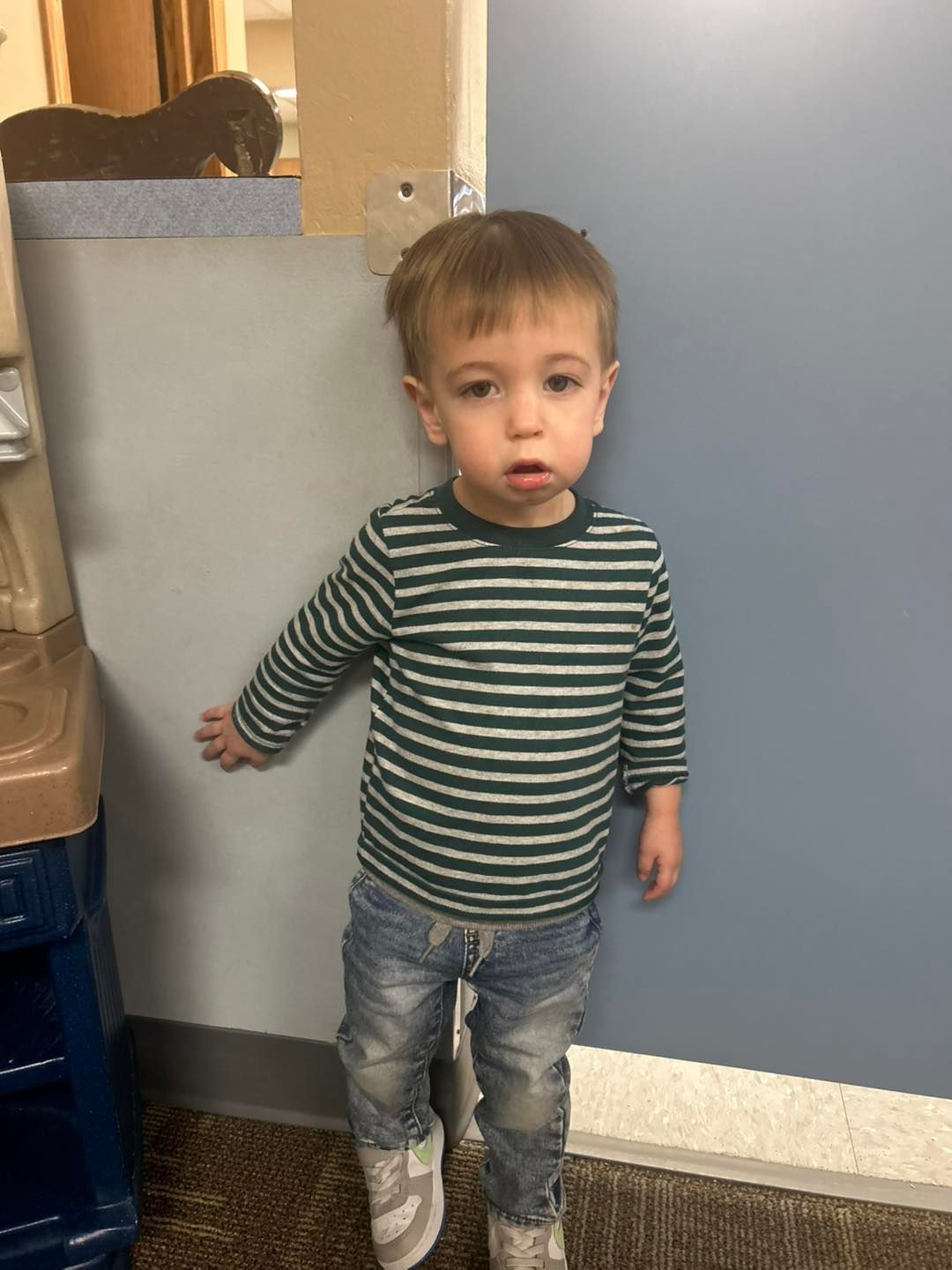 A little boy in a striped shirt and jeans is standing next to a wall.