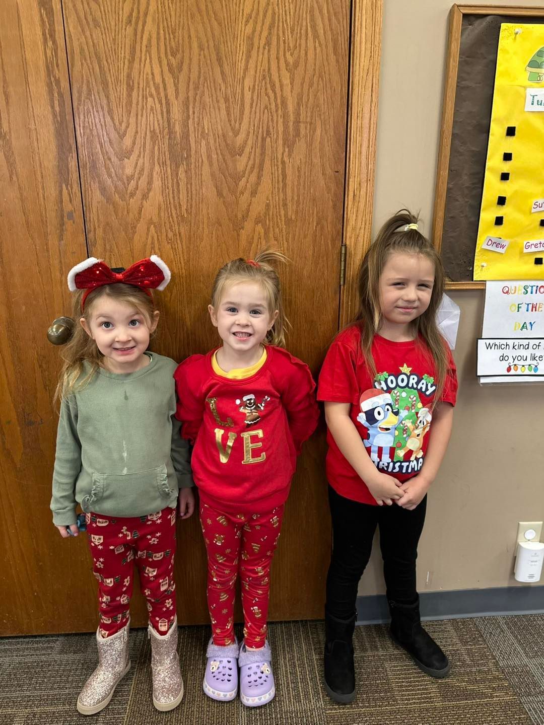 Three little girls are standing next to each other in front of a wooden door.