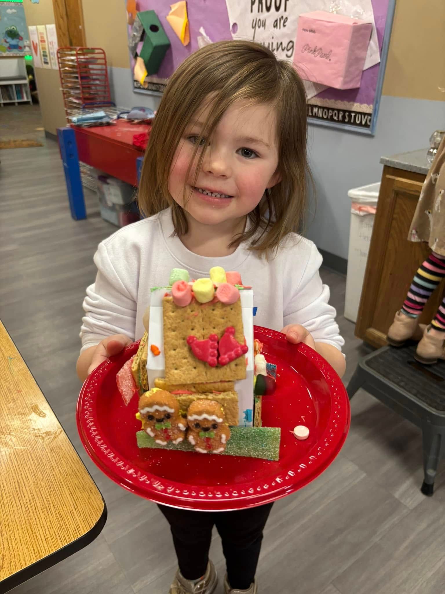 A little girl is holding a plate with a gingerbread house on it.
