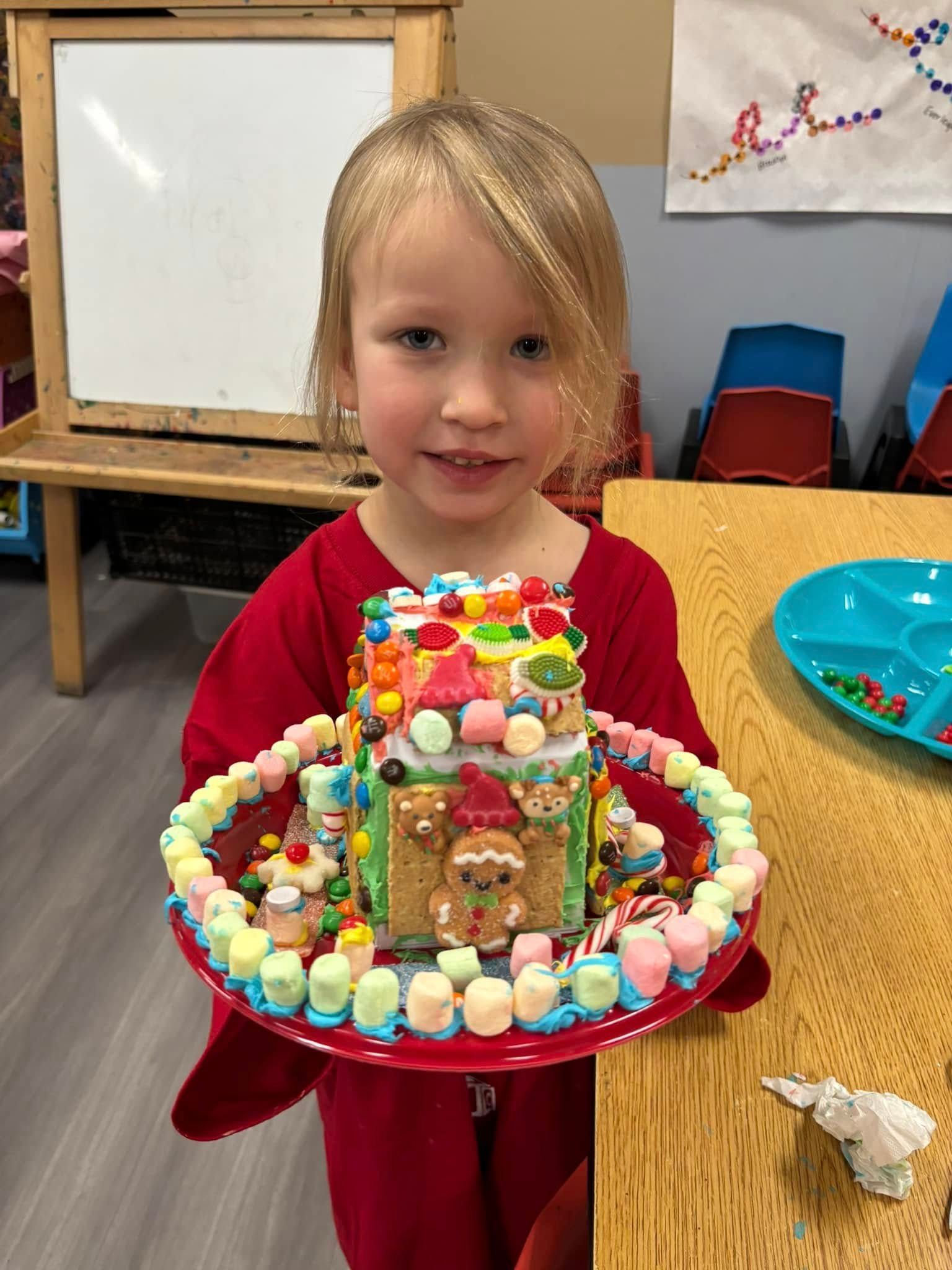 A little girl is holding a gingerbread house made of candy and marshmallows.