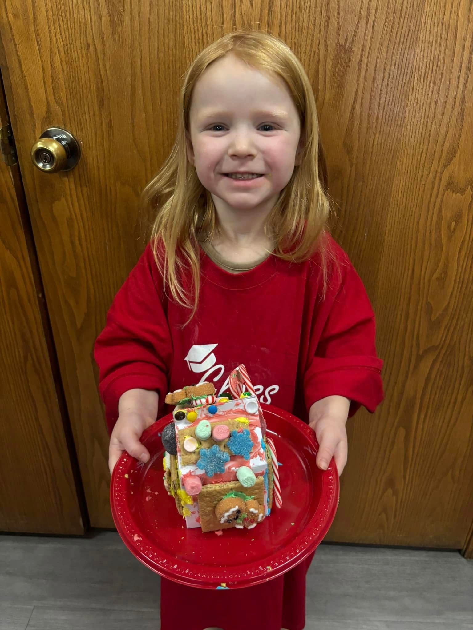 A little girl is holding a plate with a gingerbread house on it.