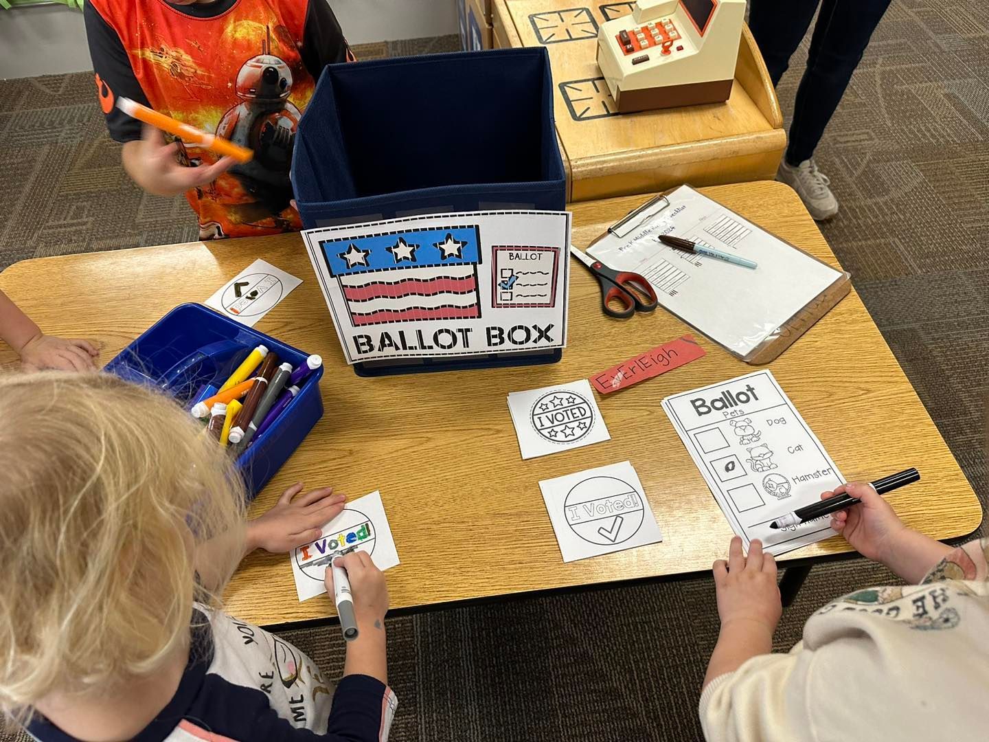 A group of children are sitting at a table with a ballot box.