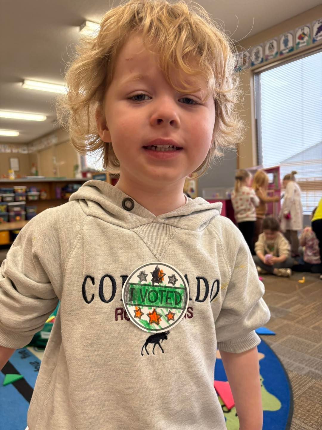 A young boy wearing a sweatshirt is standing in a classroom.