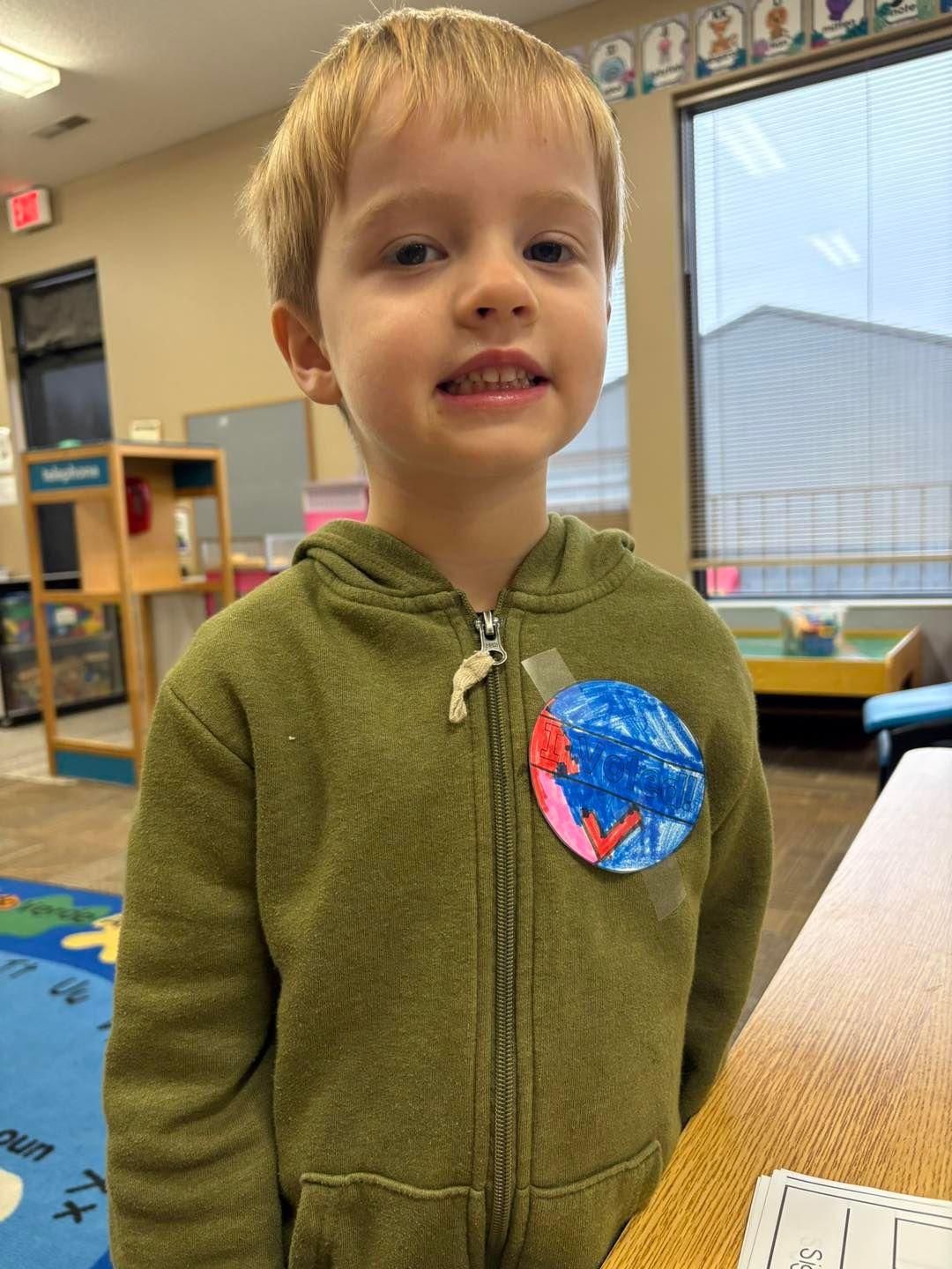 A young boy in a green jacket is standing in a classroom.