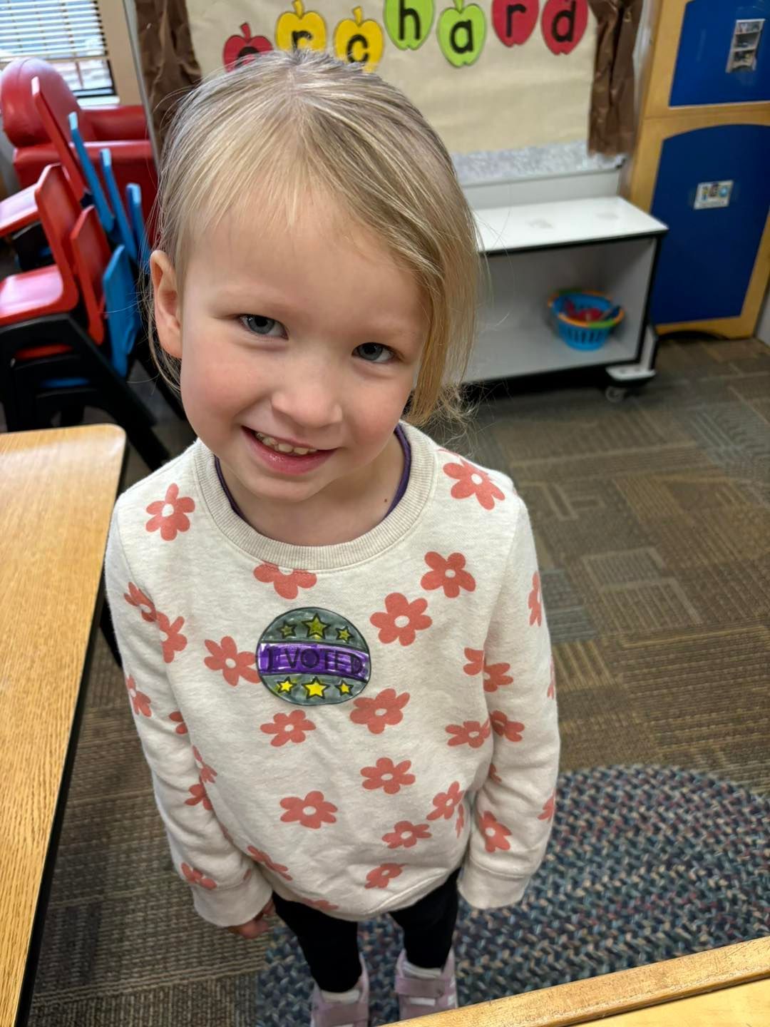 A little girl wearing a sweater with flowers on it is smiling in a classroom.